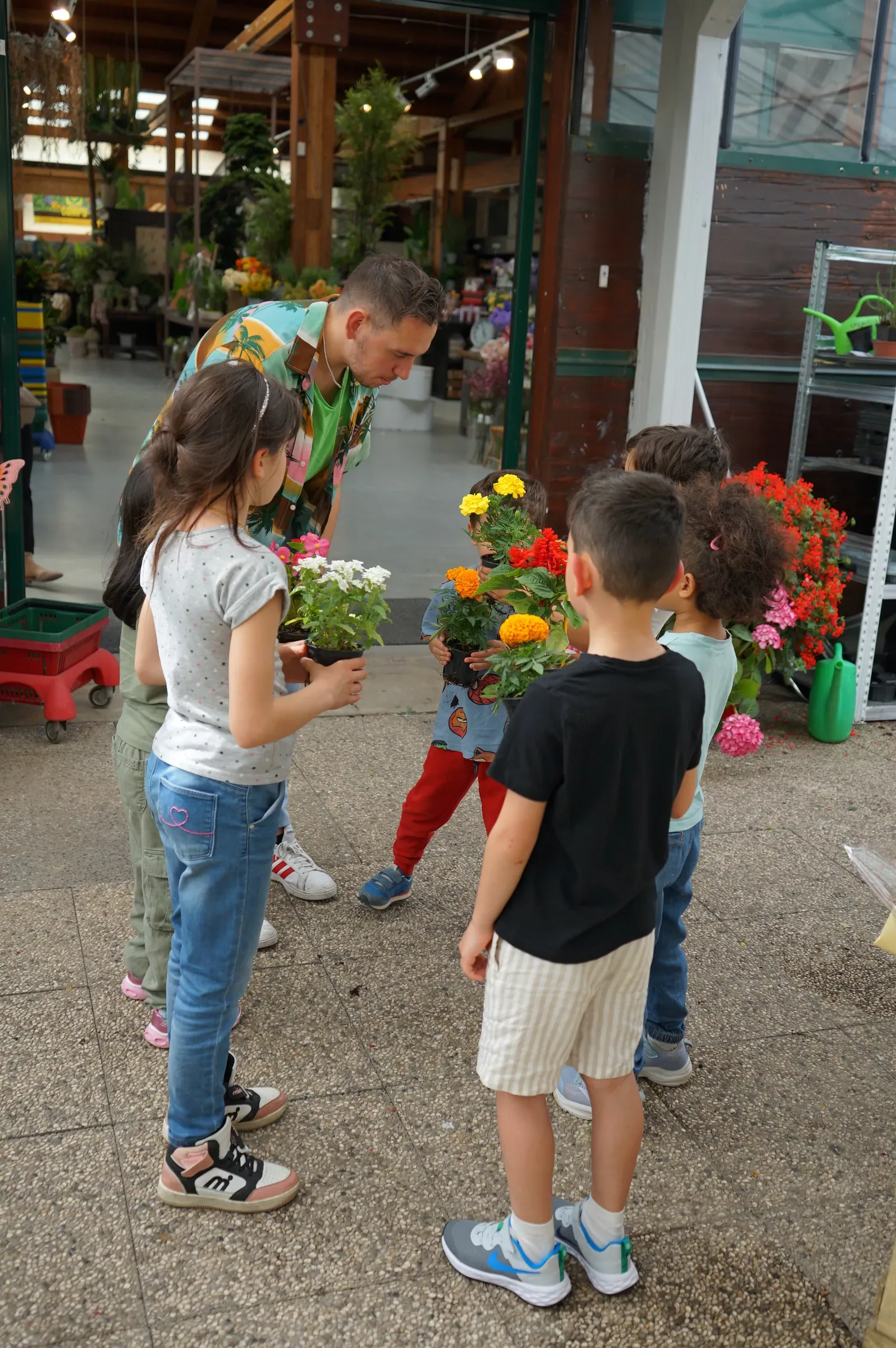 Bambini con i vasetti di fiori in mano fra i banchi del garden, con un animatore chinato verso di loro