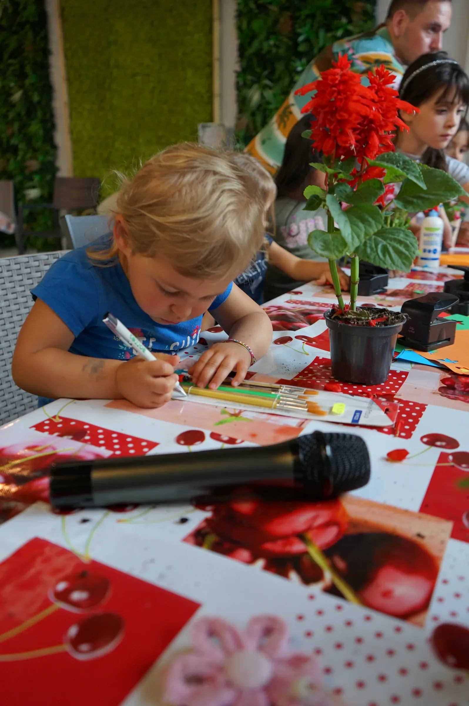 Un bambino biondo colora un cartoncino con il pennarello, al tavolo con la tovaglia a ciliegie, accanto a una salvia rossa in vaso
