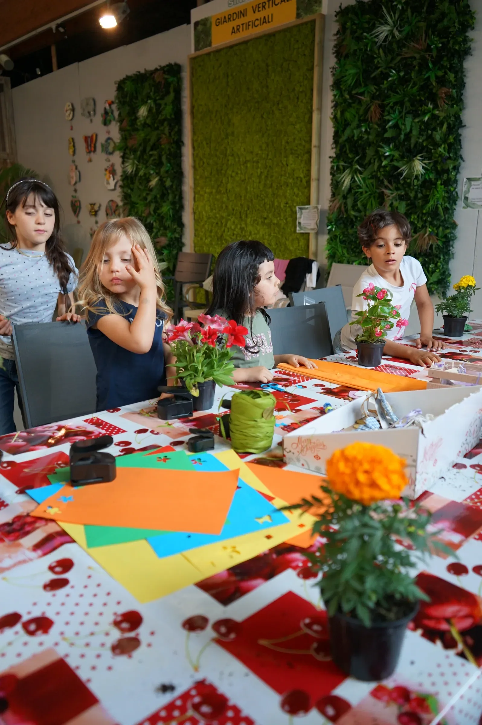 Bambine sedute al tavolo fra cartoncini colorati e vasetti di fiori, davanti alle pareti di verde