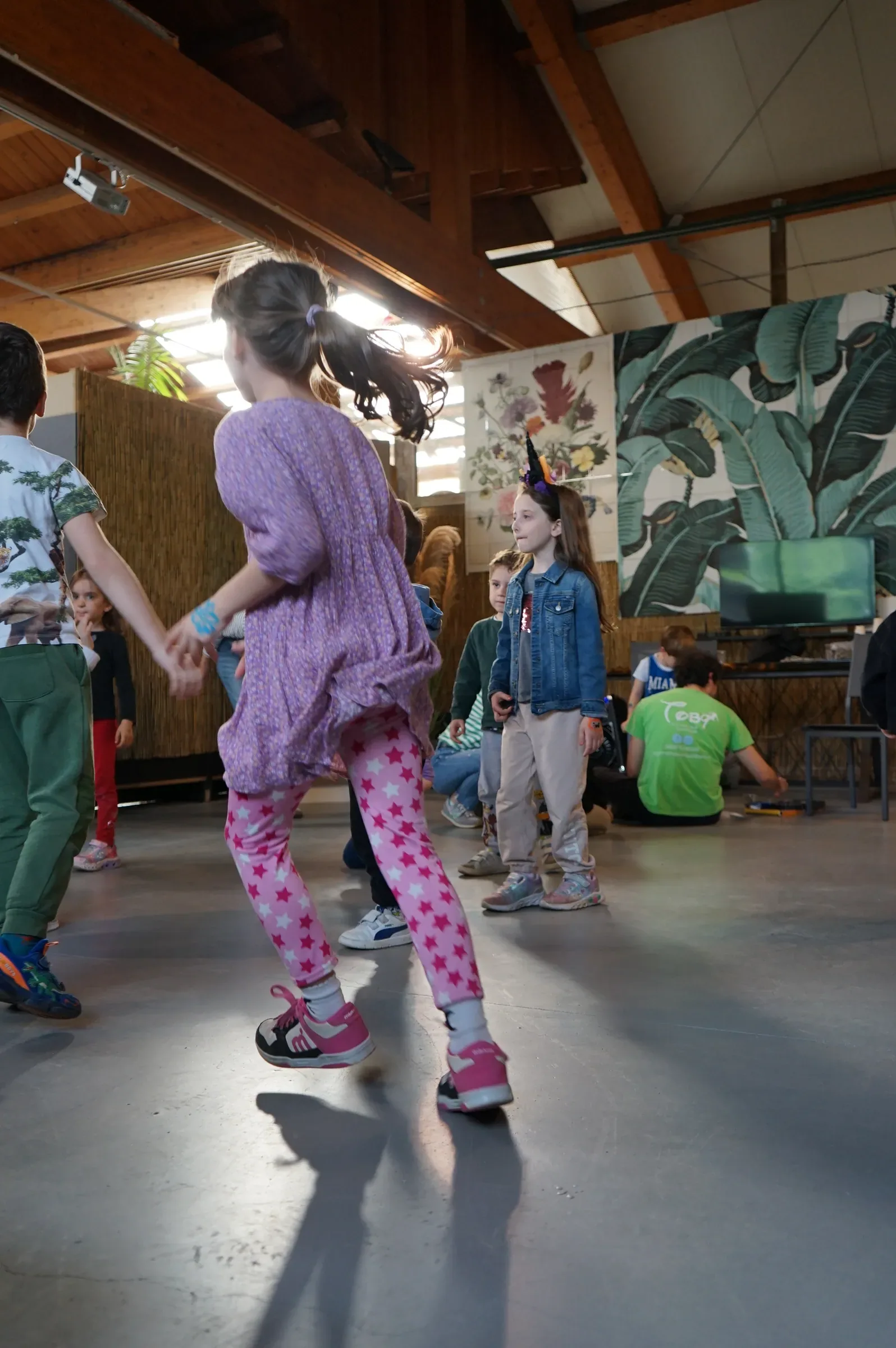 Una bambina con il vestito lilla e i leggings rosa a stelle balla sul pavimento della serra, sotto le travi di legno