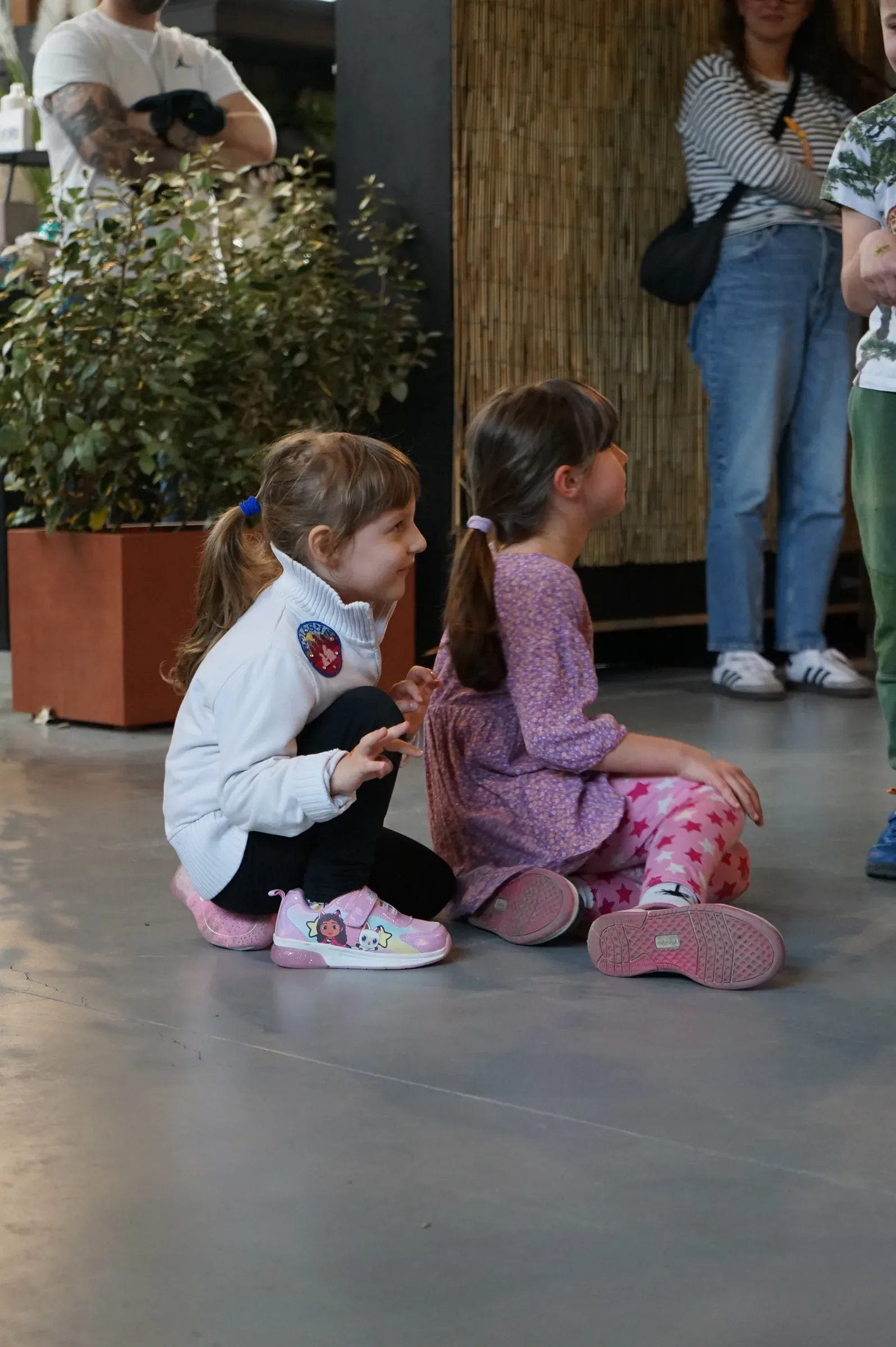 Due bambine sedute sul pavimento grigio ridono girate una verso l'altra, accanto a un grande vaso con una pianta
