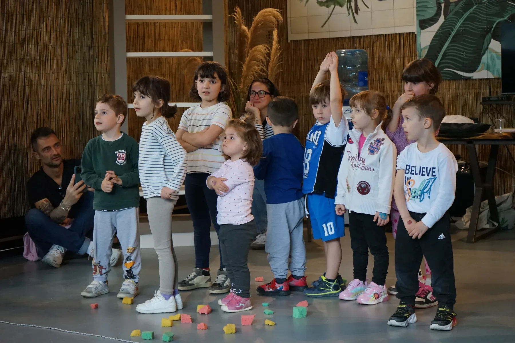Bambini in fila davanti alla parete di canne, con i cubetti colorati sparsi per terra e un adulto accovacciato a sinistra