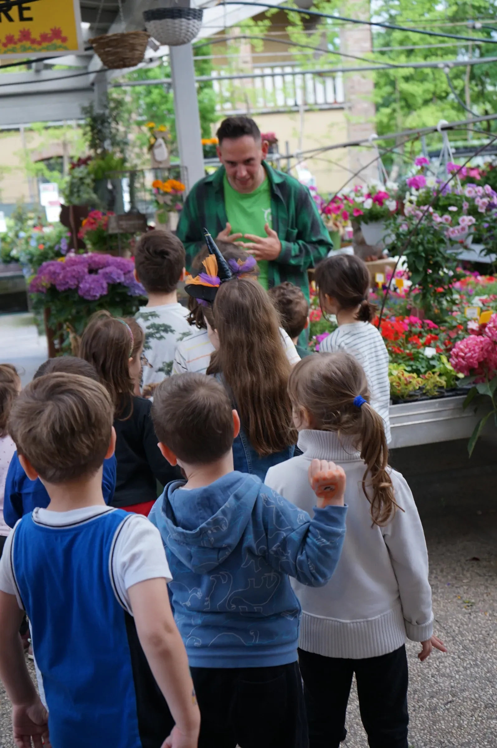 Bambini di spalle fra i banchi di fiori del garden, davanti a un animatore in maglietta verde