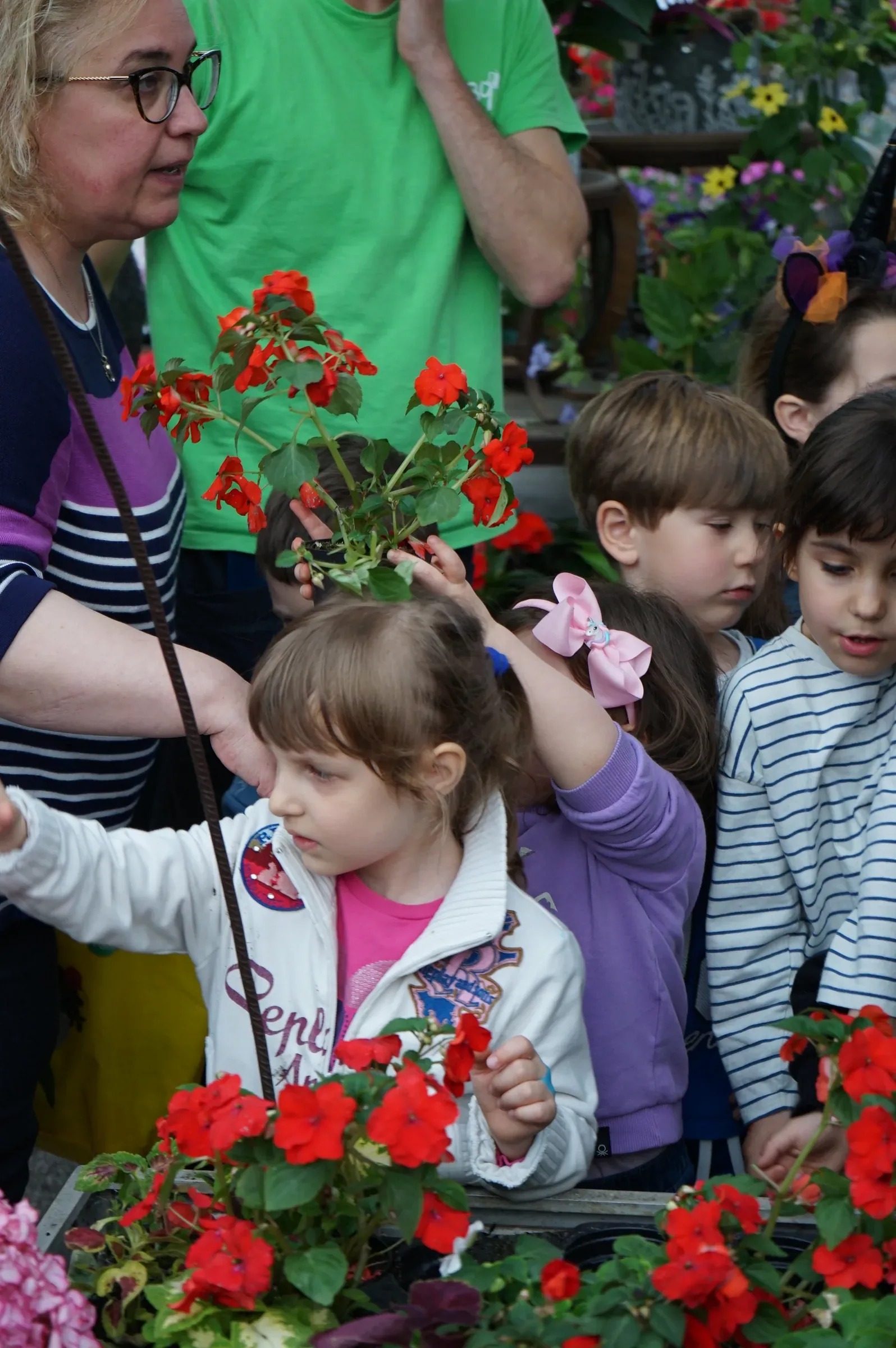 Una bambina con la felpa bianca fra altri bambini davanti a un banco di fiori rossi, con una signora con gli occhiali che indica le piante