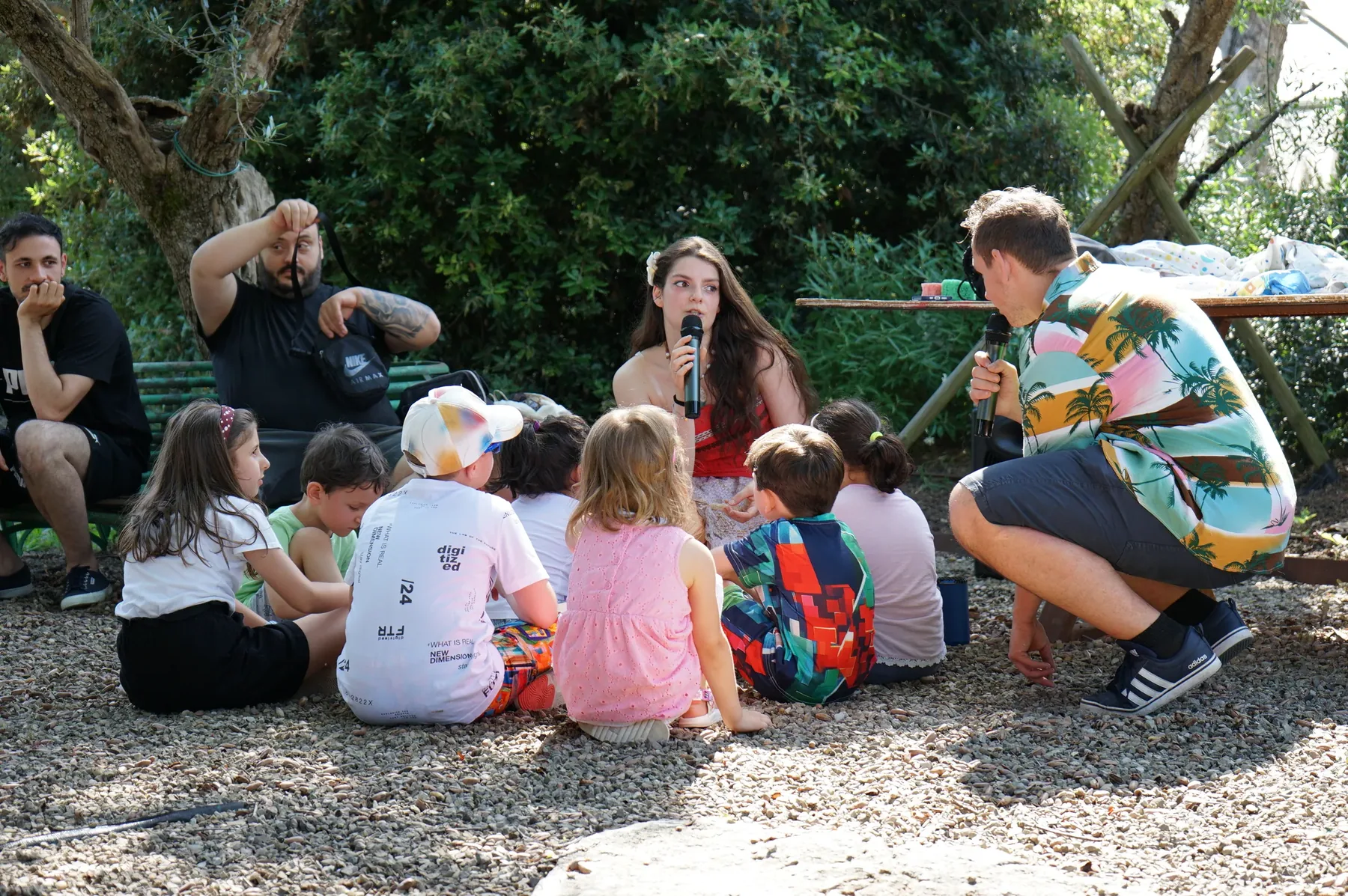 Vaiana e due animatori con il microfono seduti davanti ai bambini, sulla ghiaia all'ombra degli alberi