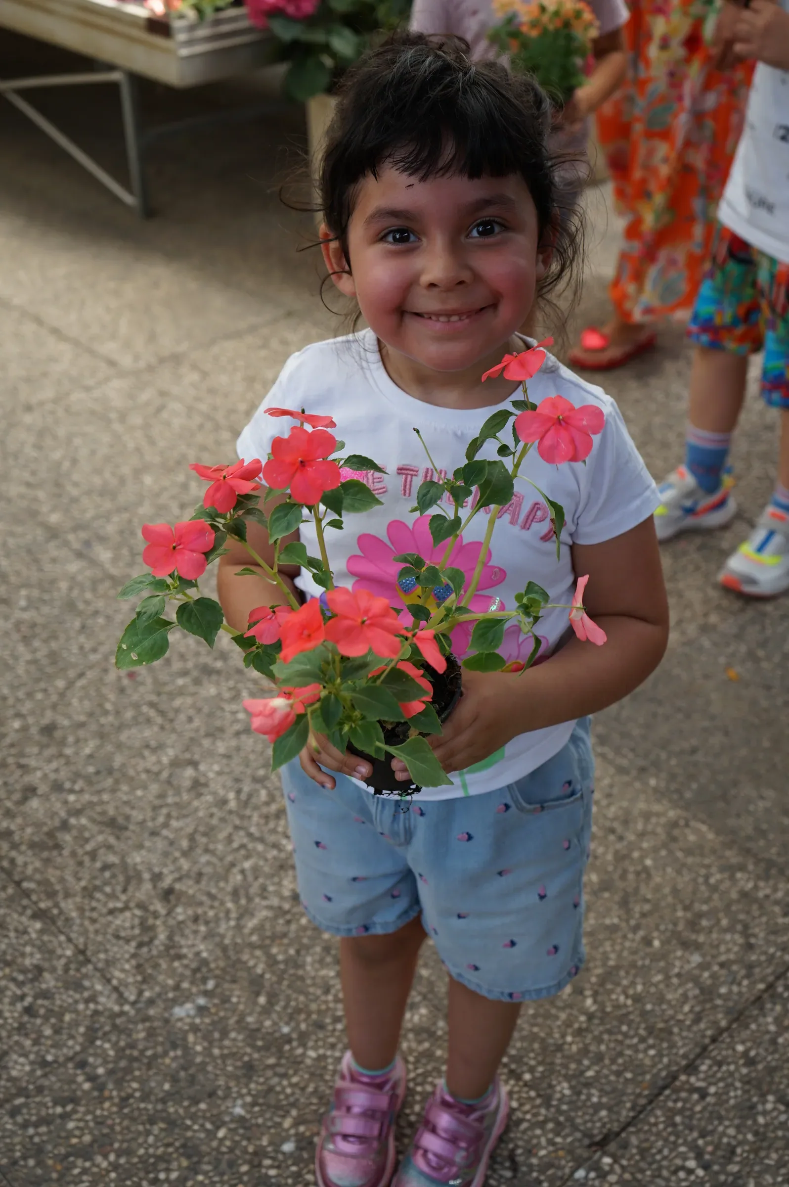 Una bambina sorride tenendo in braccio un vaso di fiori rosa corallo, nel corridoio del garden