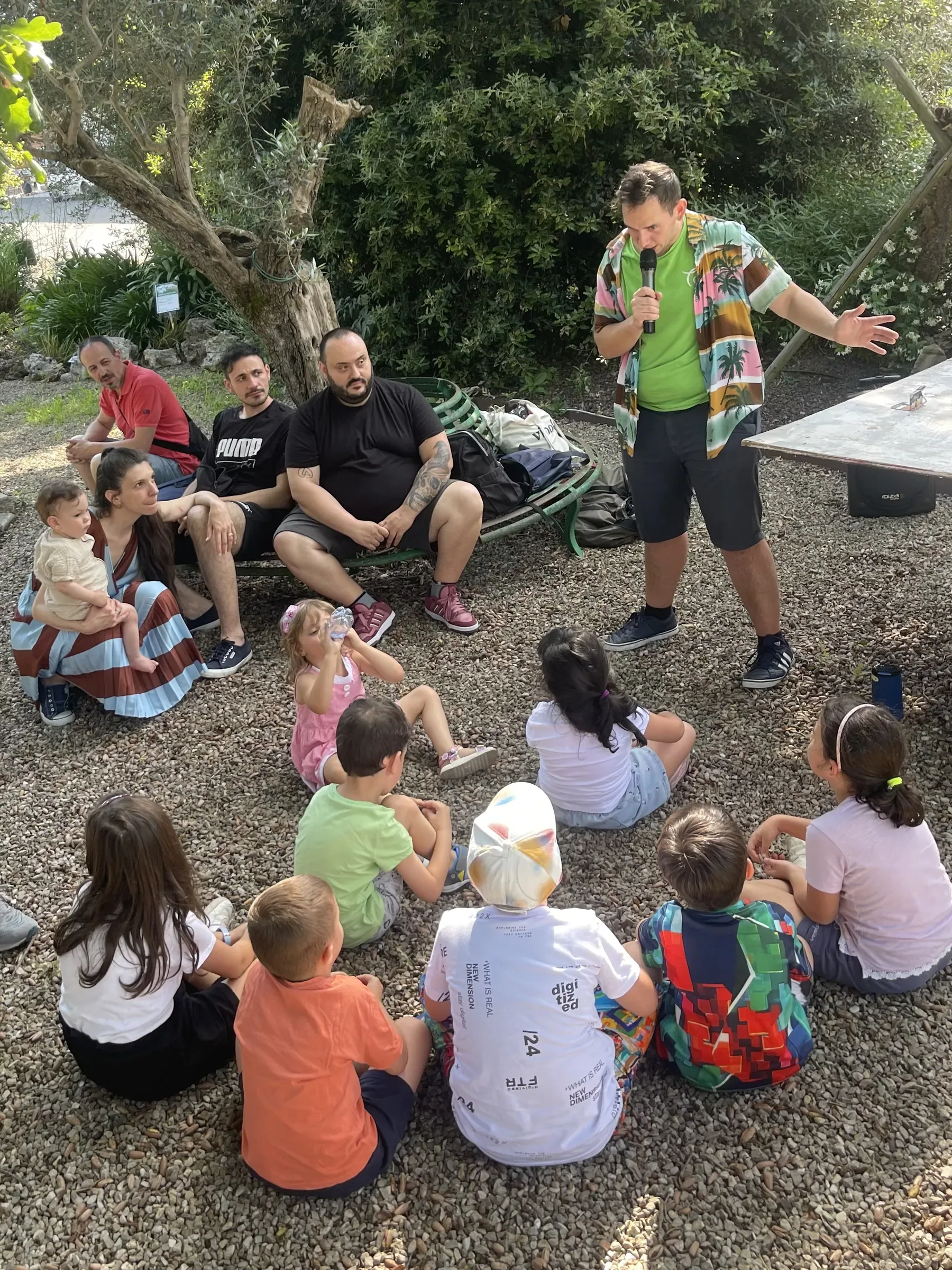 Un animatore in maglietta verde con il microfono parla ai bambini seduti sulla ghiaia, con i genitori seduti dietro