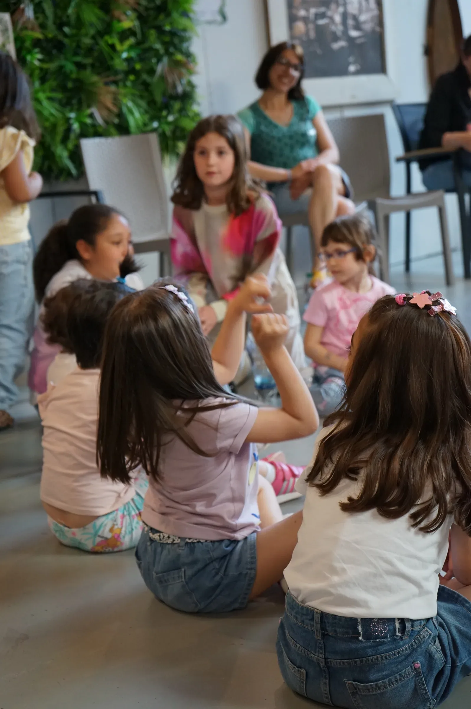 Bambine sedute in cerchio sul pavimento, alcune di spalle, con le mamme sedute dietro