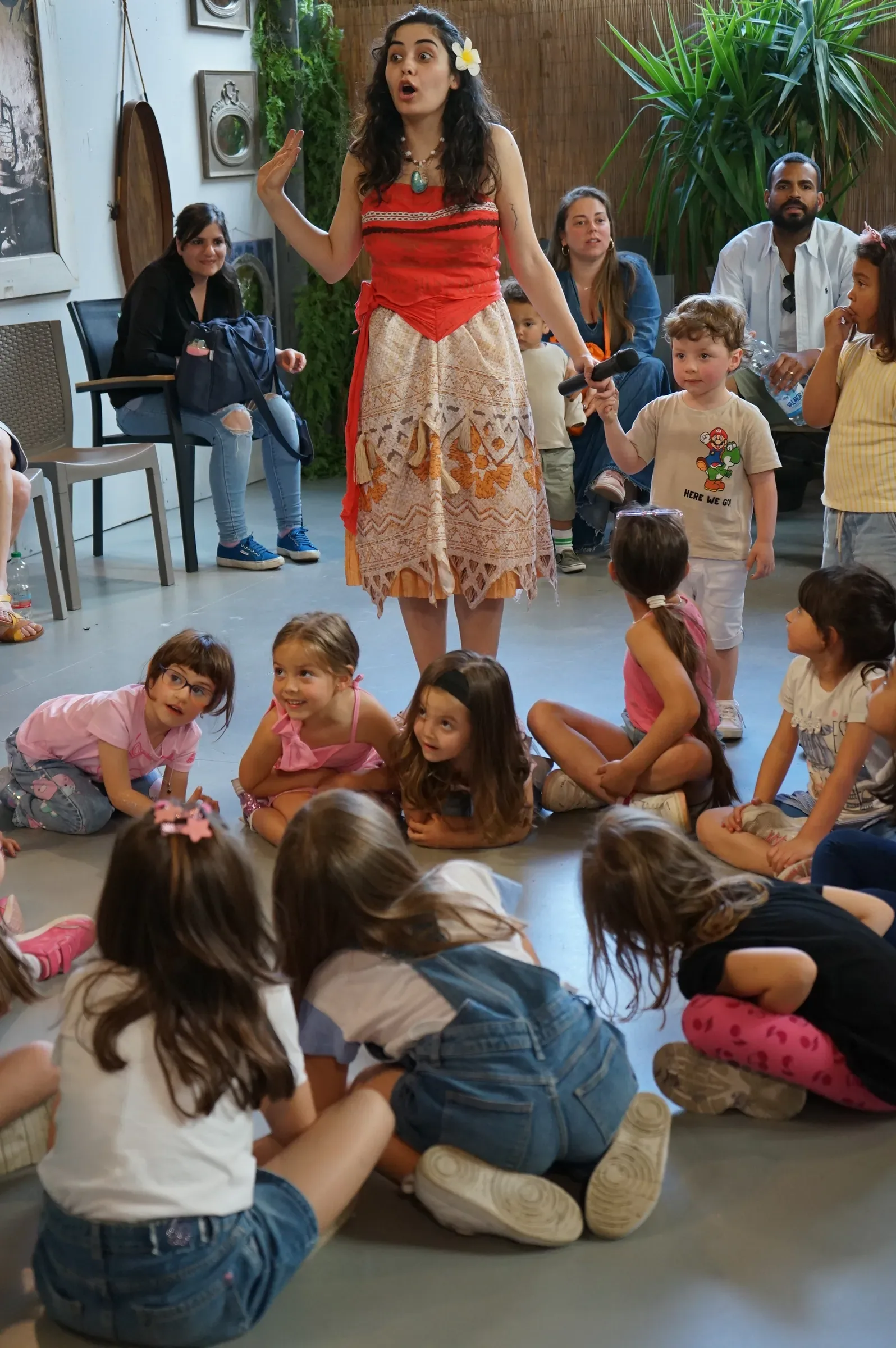 Vaiana in piedi parla ai bambini seduti in cerchio sul pavimento, con i genitori seduti dietro