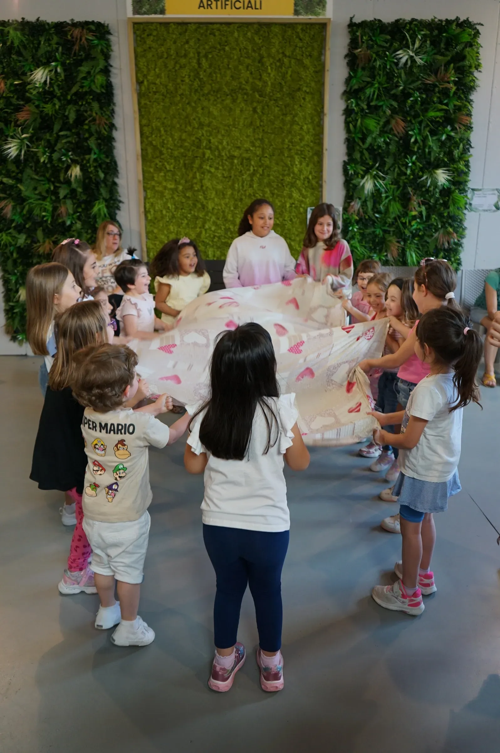 Bambini in cerchio reggono un grande telo chiaro davanti alle pareti di verde della sala