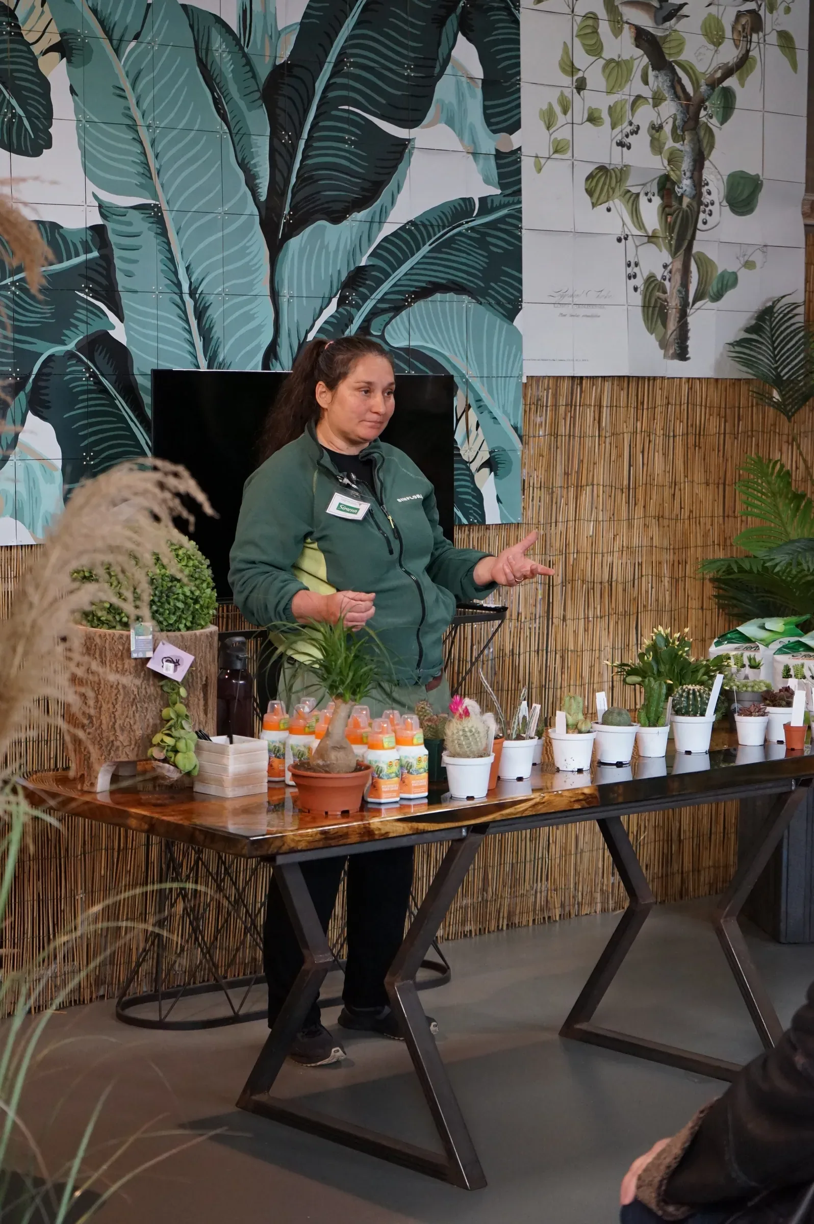 La docente in divisa verde parla dietro il tavolo lungo dei cactus, con i flaconi arancioni