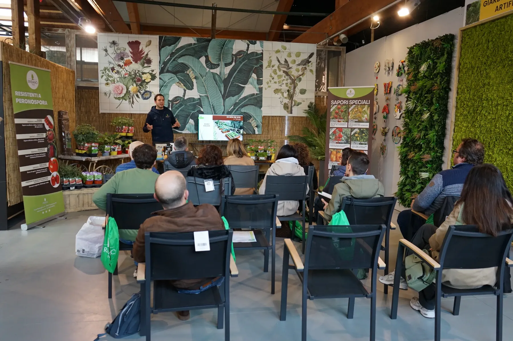 Il relatore parla davanti allo schermo e i partecipanti lo ascoltano seduti, fra gli espositori delle piantine da orto