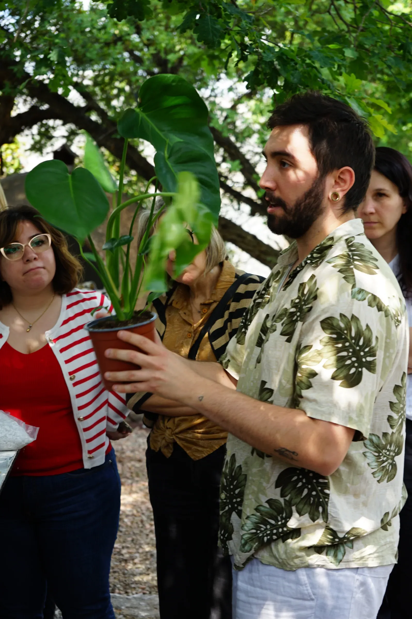 Un ragazzo con la camicia a foglie tiene una monstera nel vaso, fra le partecipanti attorno al tavolo in giardino