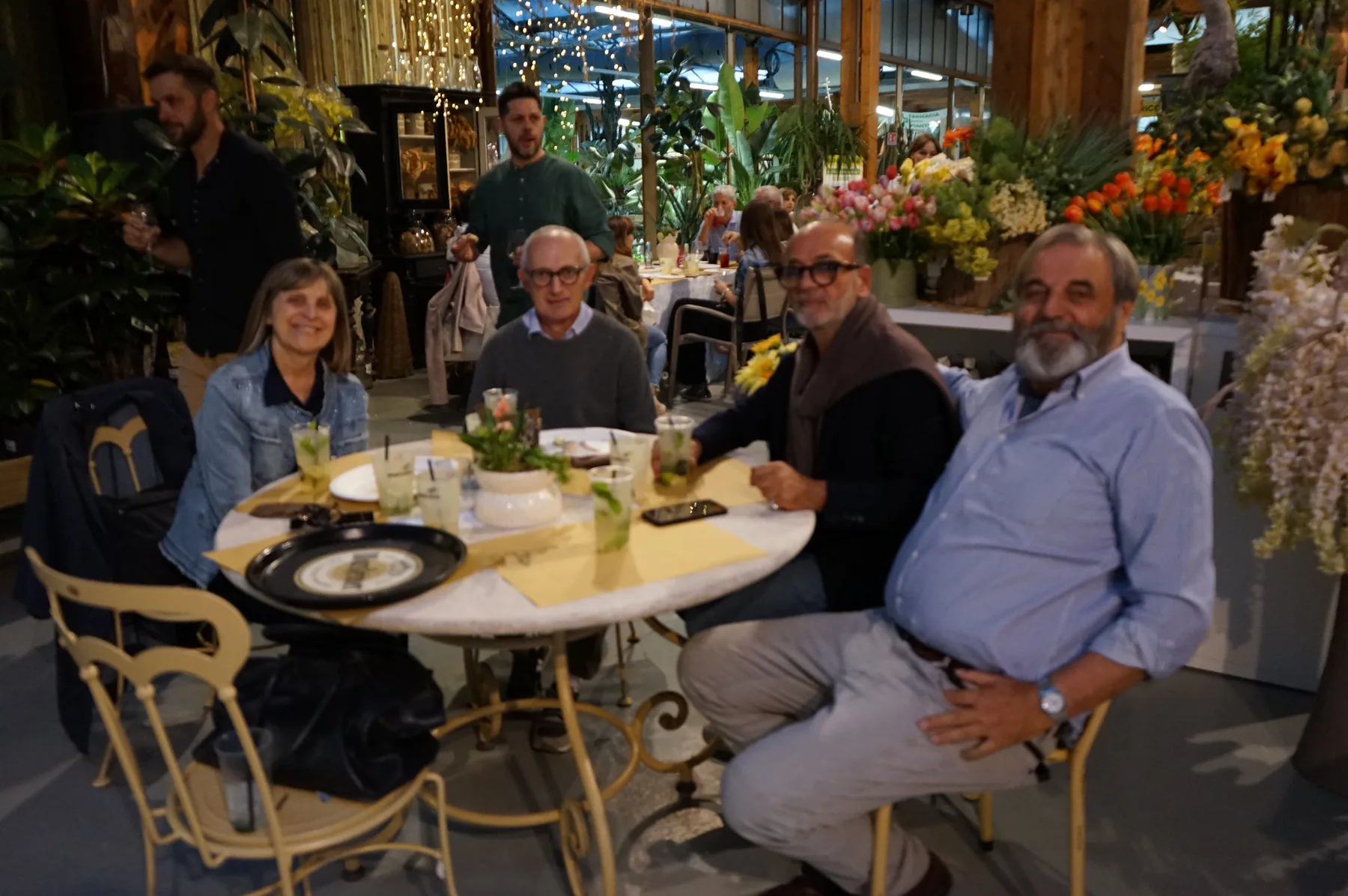 Quattro ospiti sorridenti intorno a un tavolo rotondo con i bicchieri e una piantina fiorita, con i fiori della fioreria alle spalle
