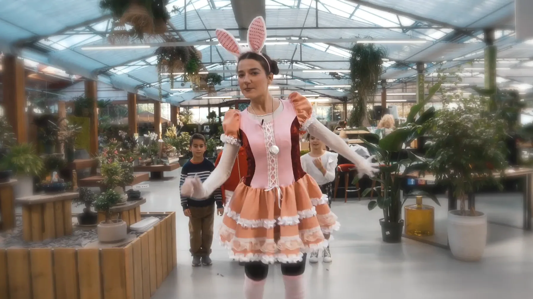 Un'animatrice vestita da coniglio, con le orecchie rosa e il vestito a balze, balla fra i banchi di piante della serra con due bambini dietro