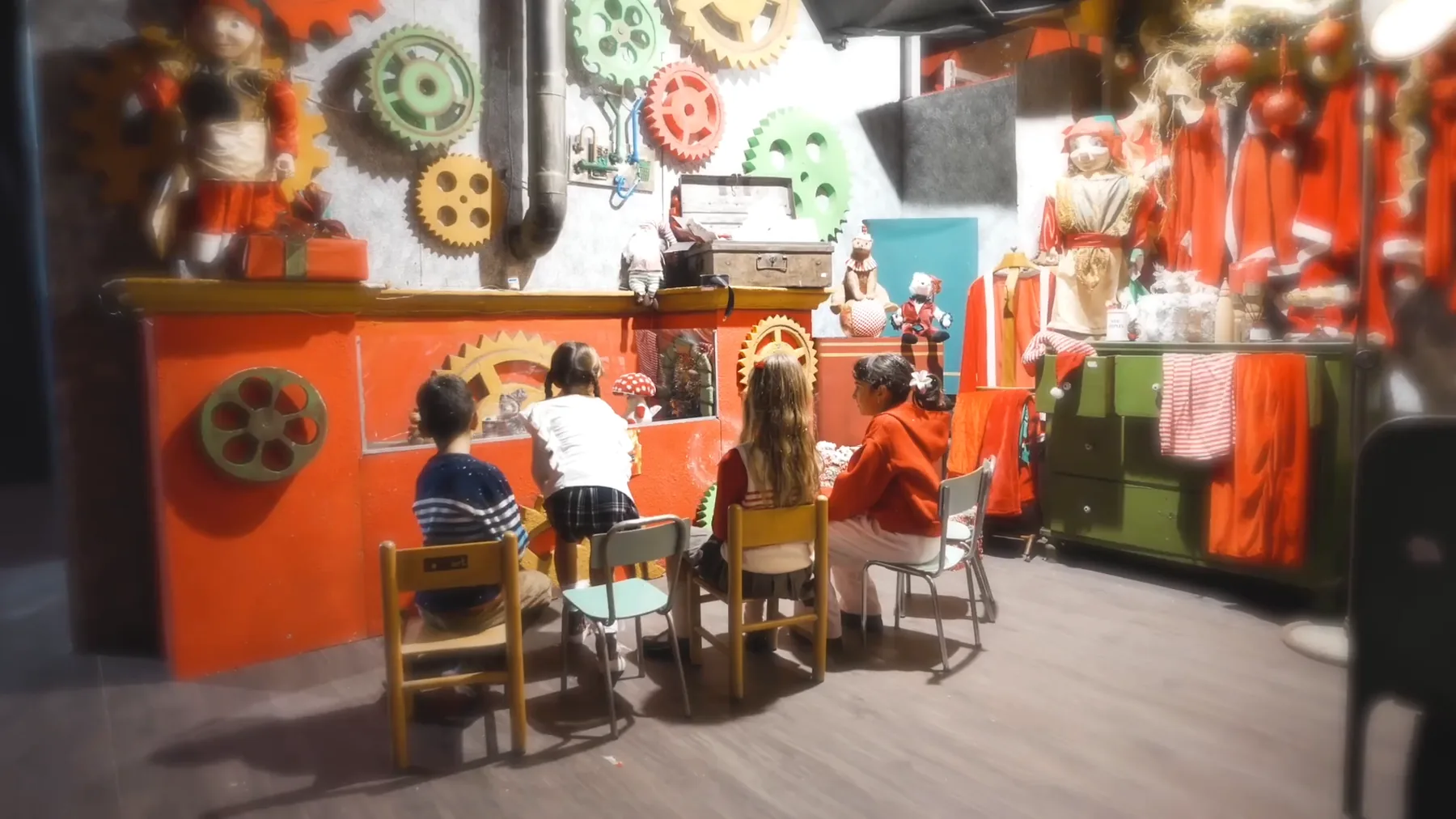 Quattro bambini seduti ai banchetti del laboratorio degli elfi, fra ingranaggi colorati e costumi rossi appesi