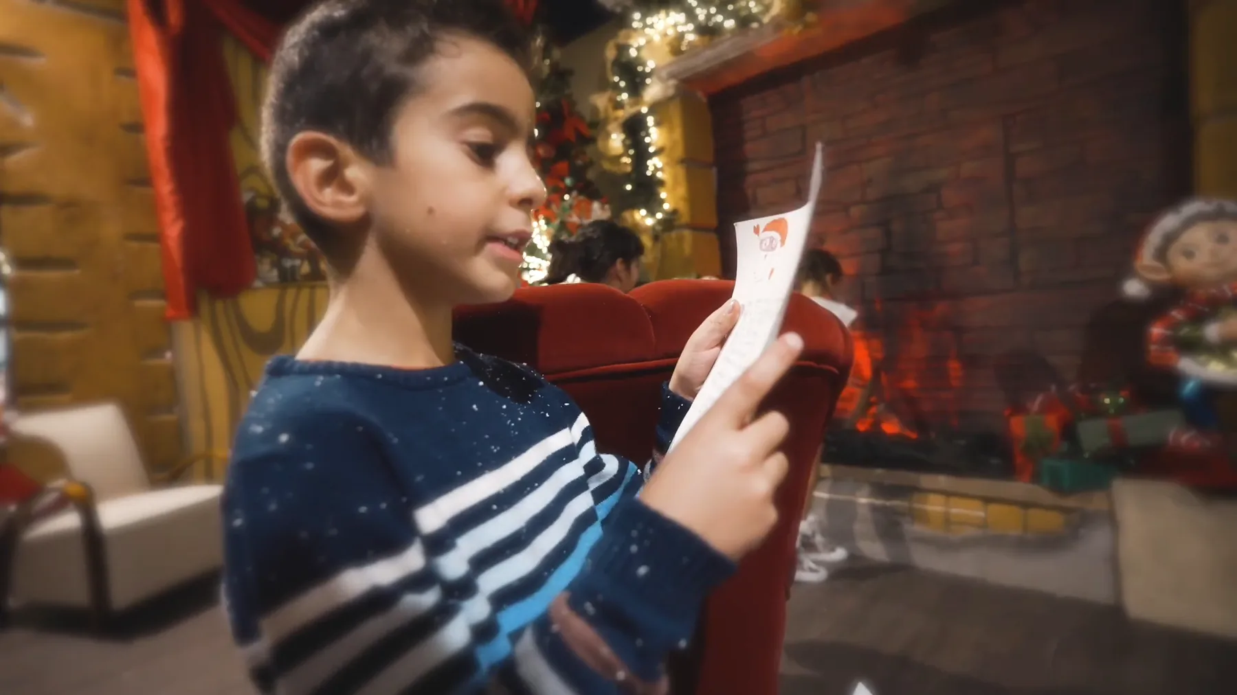 Un bambino con il maglione a righe legge una letterina davanti al camino della casa di Babbo Natale