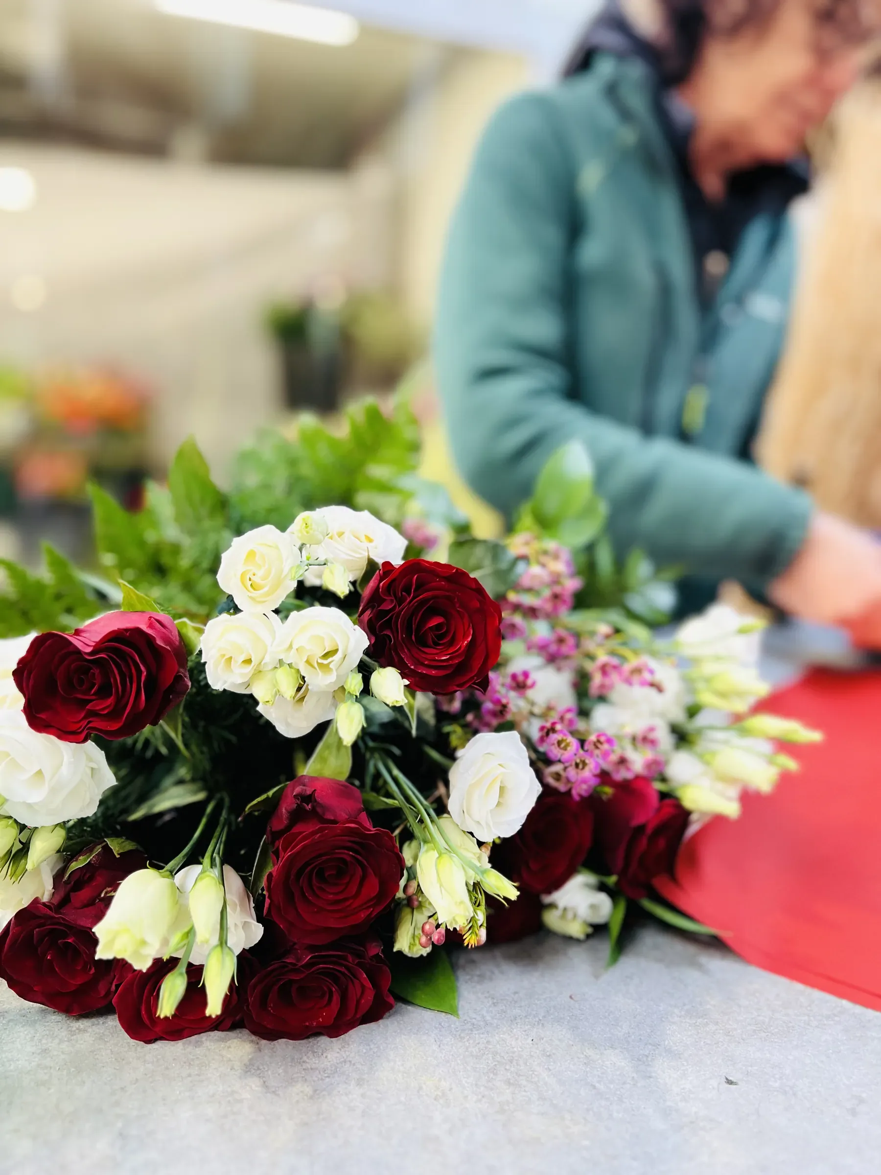 Rose rosse, lisianthus bianchi e fiorellini rosa distesi sul banco e, sfocata dietro, la fiorista al lavoro sulla carta rossa