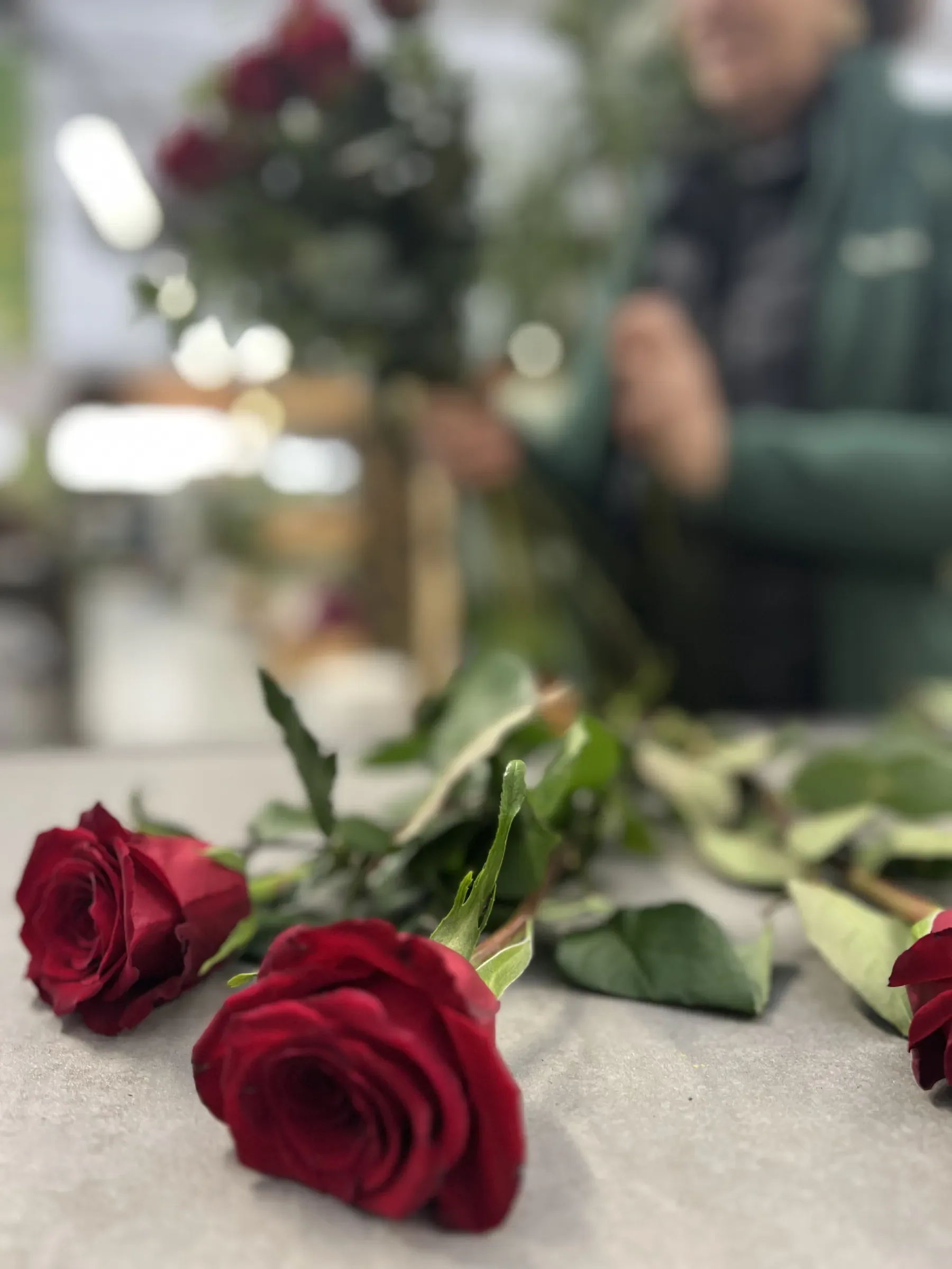 Due rose rosse distese sul banco e, sfocata dietro, la fiorista che lega un mazzo di rose rosse