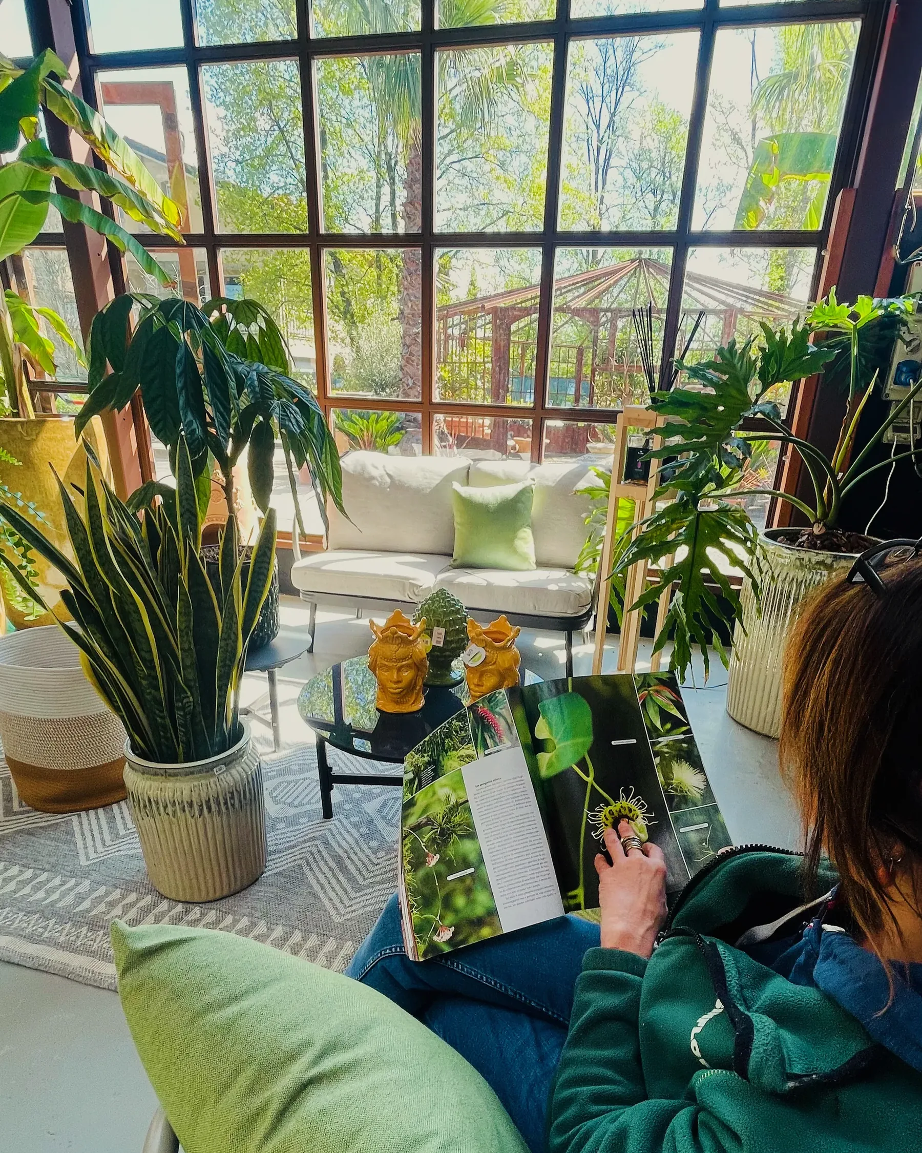 Una donna seduta su un cuscino verde sfoglia un libro di piante, nella veranda con il divano chiaro, le teste di moro gialle sul tavolino e le vetrate a quadri