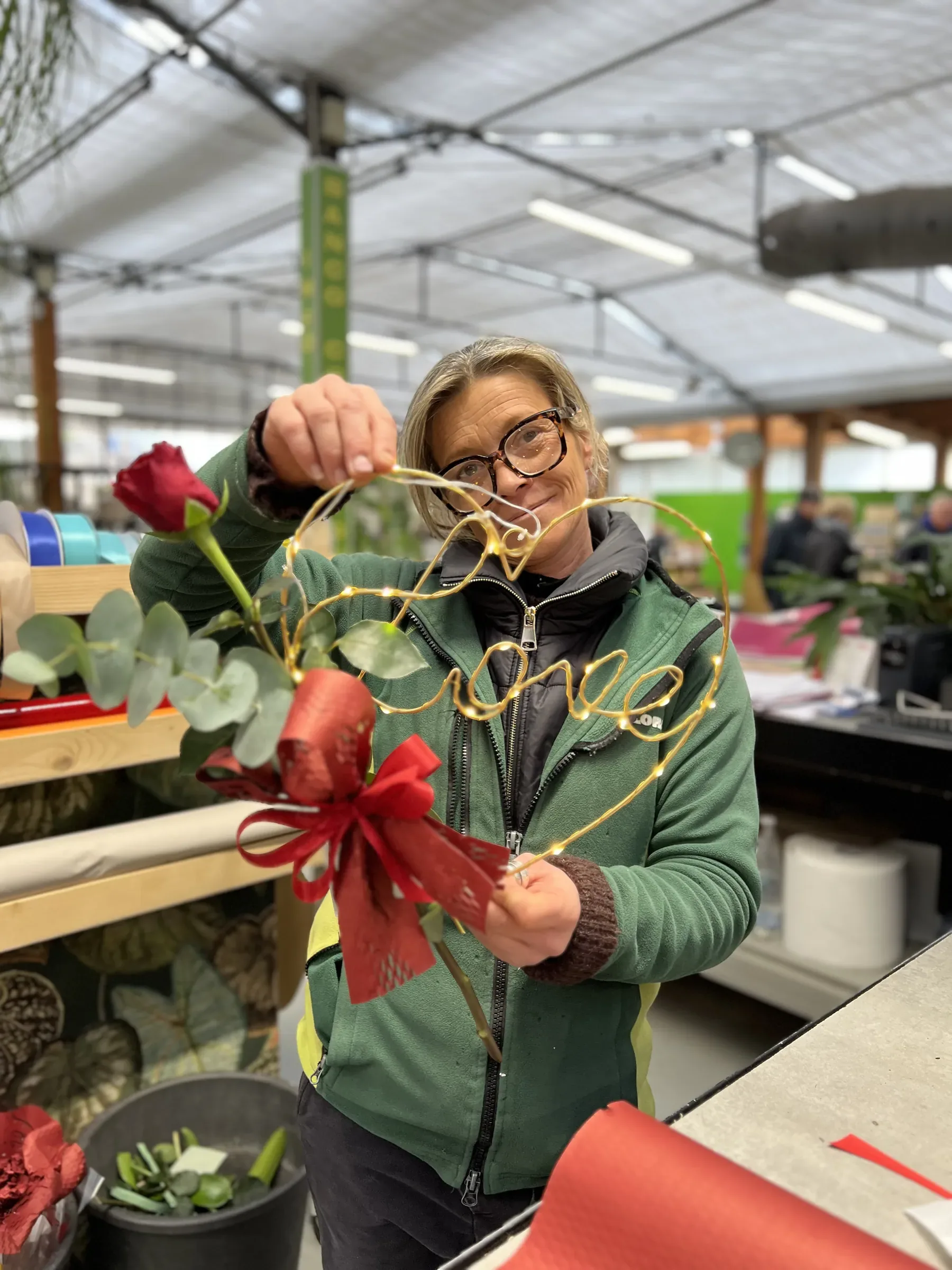 Al banco sotto la serra, una fiorista in giacca verde sorride mentre lega un fiocco rosso a una rosa con l'eucalipto e una scritta in filo dorato