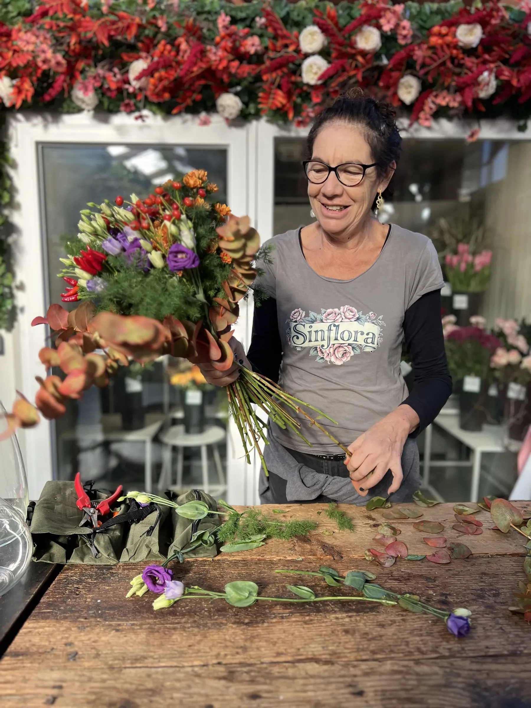 Una fiorista con la maglietta Sinflora sorride al banco di legno mentre compone un mazzo di fiori misti ed eucalipto, sotto una fascia di fiori rossi e bianchi