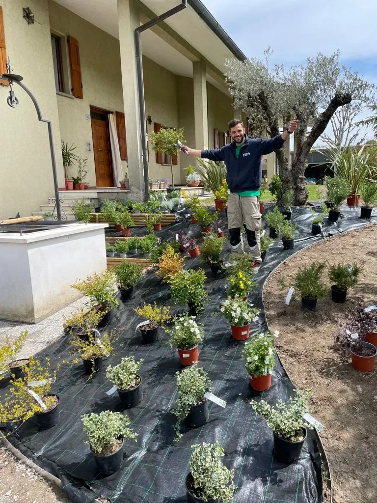Piante in vaso disposte sul telo di una nuova aiuola dal bordo curvo, pronte per la messa a dimora, con un giardiniere sorridente in mezzo e una casa sul fondo
