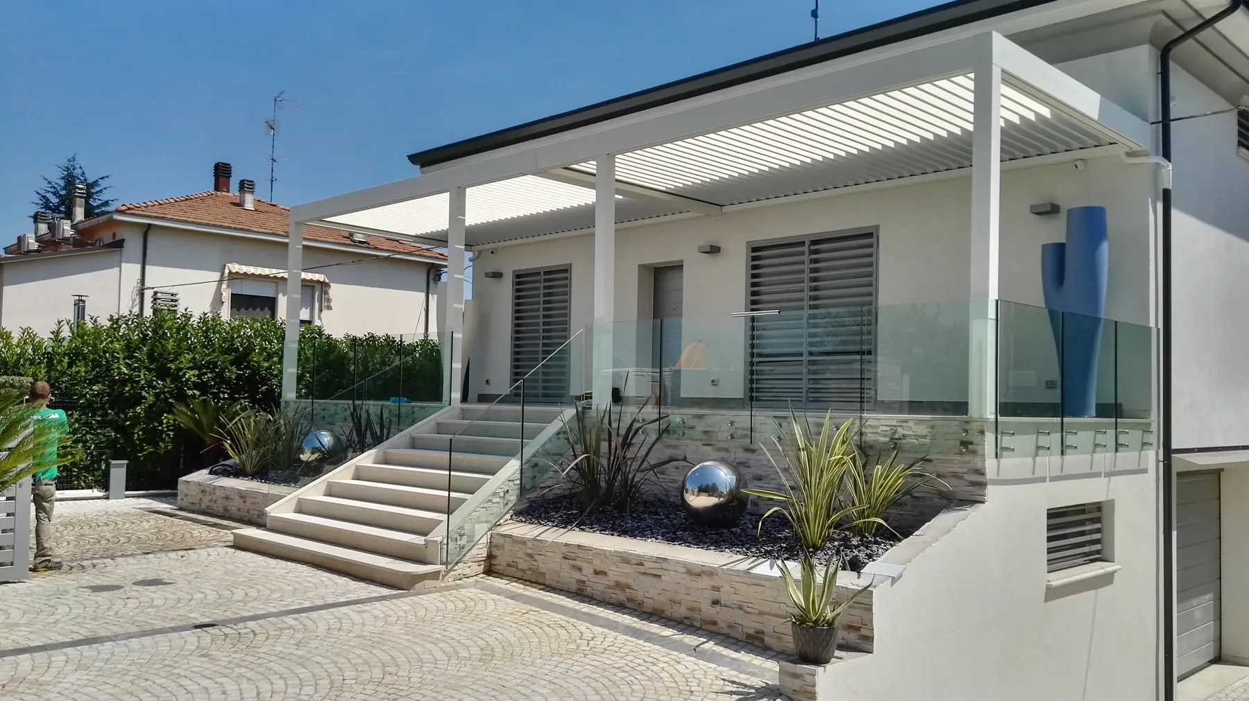Pergola bianca a lamelle sopra l'ingresso di una casa, con scalinata chiara, parapetto in vetro e aiuole di ardesia con sfere d'acciaio