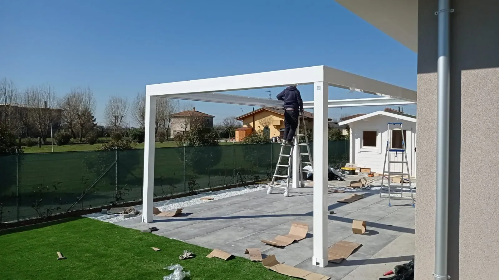 Operaio su una scala che monta la copertura di una pergola bianca, sopra un pavimento in lastre grigie accanto al prato