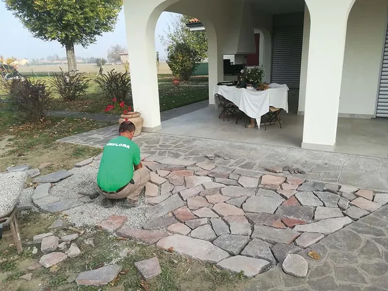 Giardiniere in maglietta verde che posa lastre di pietra irregolari su un letto di ghiaia, davanti a un portico ad archi