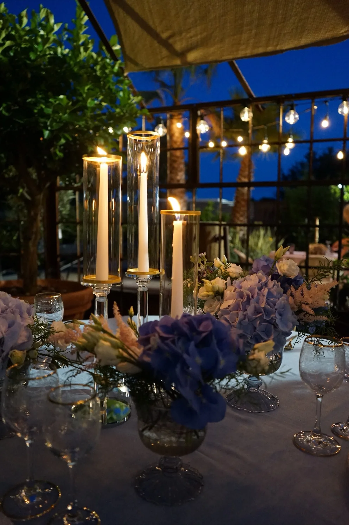 Candele accese nei cilindri di vetro fra ortensie blu e fiori chiari, di sera