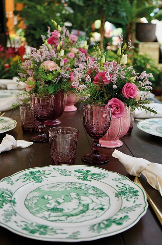 Piatto bianco a decoro verde, calici rosa e composizioni di rose e fiori rosa su un tavolo di legno in serra
