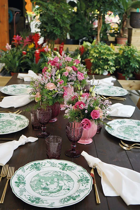 Tavolo lungo apparecchiato con piatti a decoro verde, tovaglioli bianchi e vasi di fiori rosa