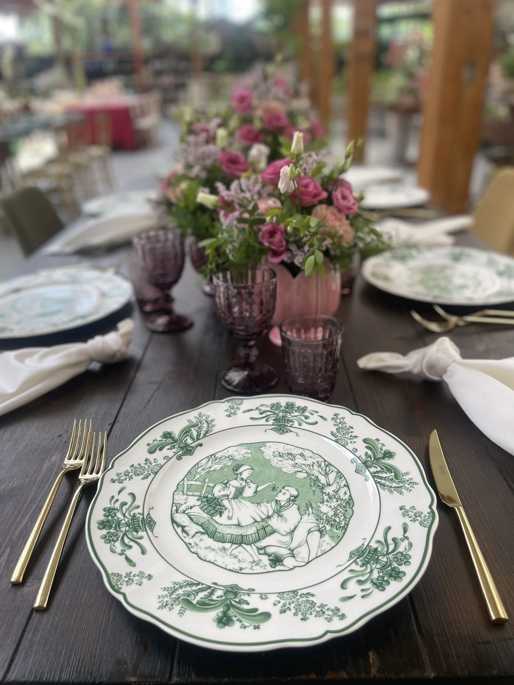 Piatto di ceramica a decoro verde con le posate dorate e i calici rosa sullo sfondo
