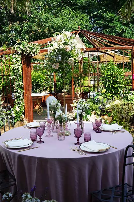 Tavola rotonda rosa antico con calici viola in giardino, davanti al pergolato fiorito