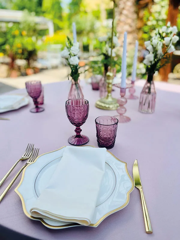 Coperto con piatto bordato d'oro, calice viola e vasetto di fiori bianchi sulla tovaglia rosa