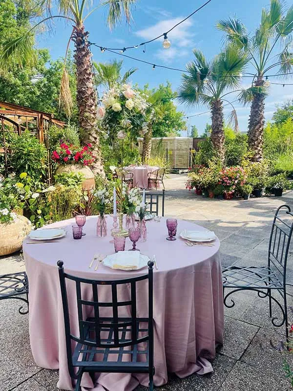 Tavola rosa in giardino fra palme e fiori, con le lucine appese