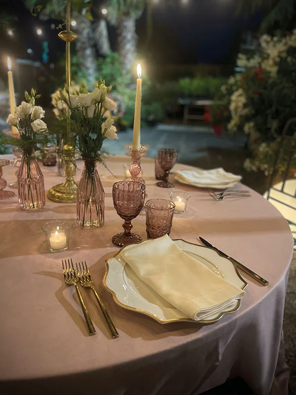 Candele accese, vasetti di fiori chiari e calici viola sulla tavola rosa, di sera